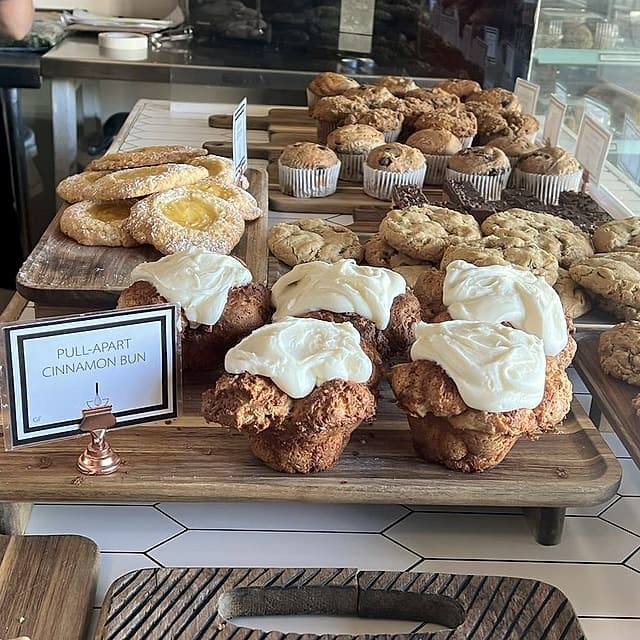 Frosted pull-apart cinnamon buns on a wooden tray, with scones and muffins behind