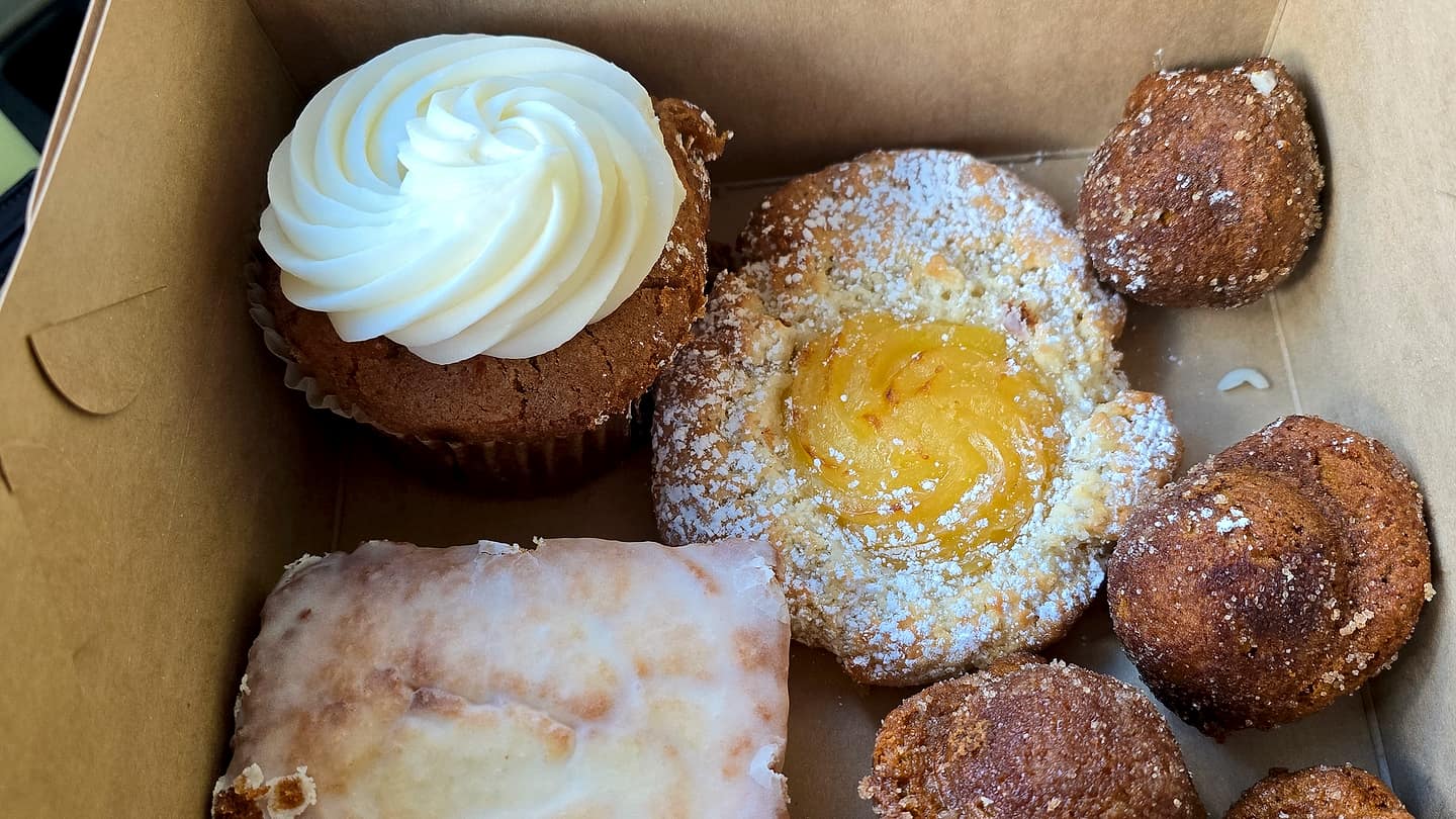 A box of pastries: a cupcake with a swirl of white frosting, a lemon tart dusted with powdered sugar, sugared donut holes, a glazed bar and a chocolate bar topped with cacao nibs