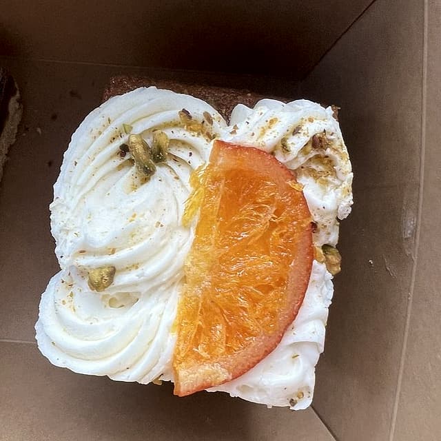 A square of cake with swirled white frosting, a candied orange slice and chopped pistachios